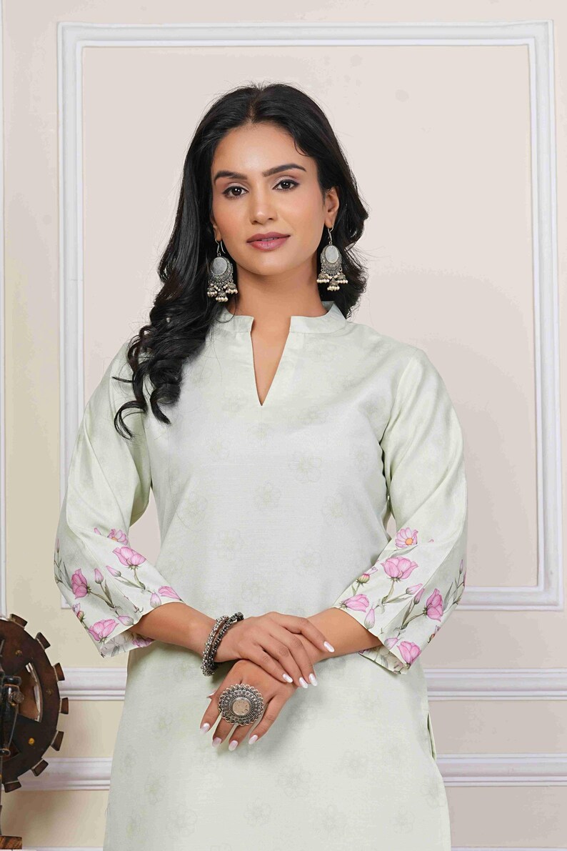 May include: A light green tunic with a floral pattern and a mandarin collar. The sleeves are adorned with pink floral designs. The model is wearing silver earrings, a bracelet, and a large ring. This outfit is suitable for casual or semi-formal occasions.