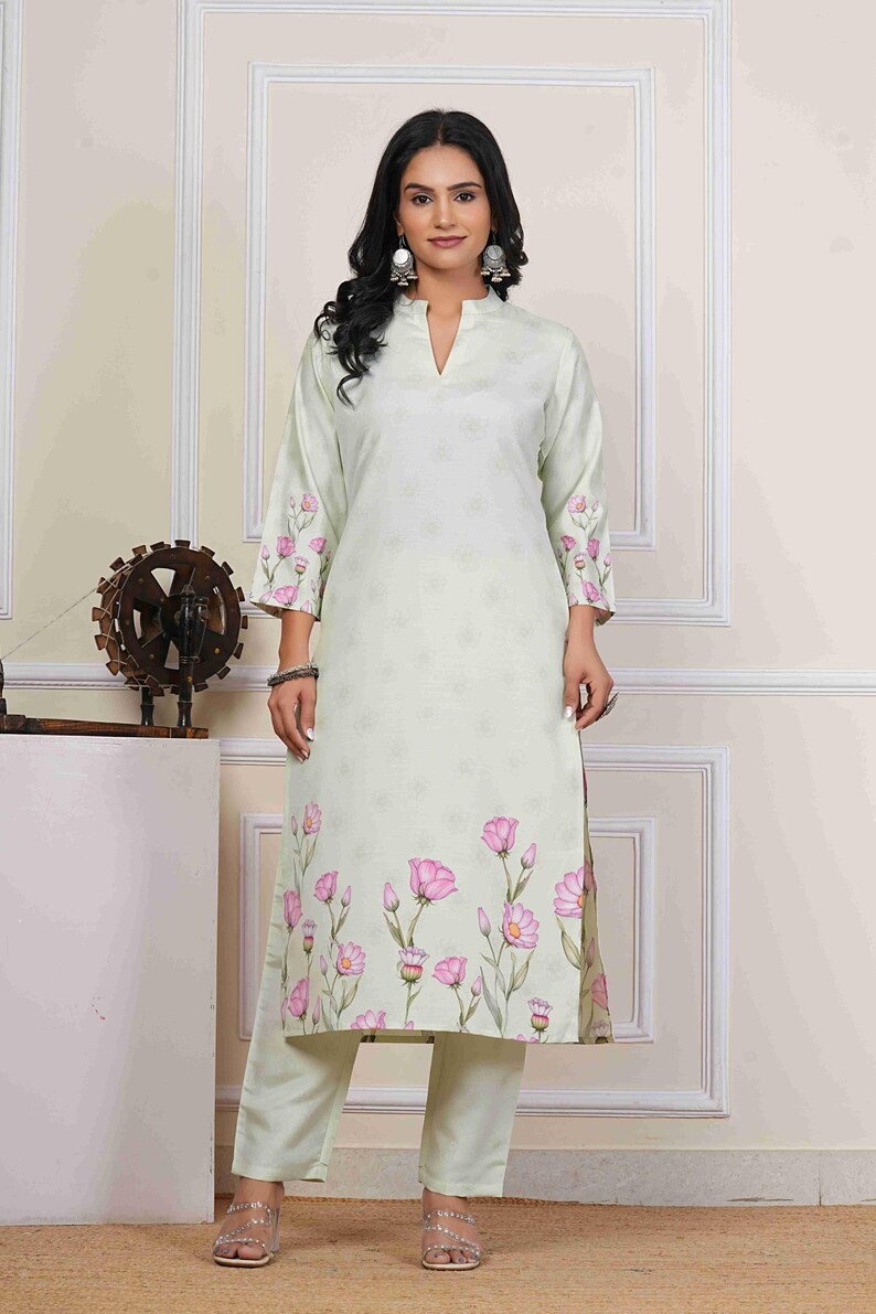 May include: A light green floral print kurta and matching pants set. The kurta features a V-neck and three-quarter sleeves with pink floral accents. The floral pattern continues along the bottom of the kurta. The pants are a solid light green color.