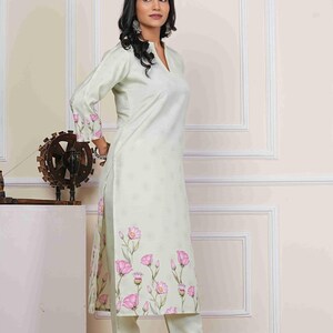 May include: A light green, long-sleeved kurta and matching pants set. The kurta features a V-neck and is adorned with a pink floral print along the hem and sleeves. The outfit is made of a lightweight fabric.