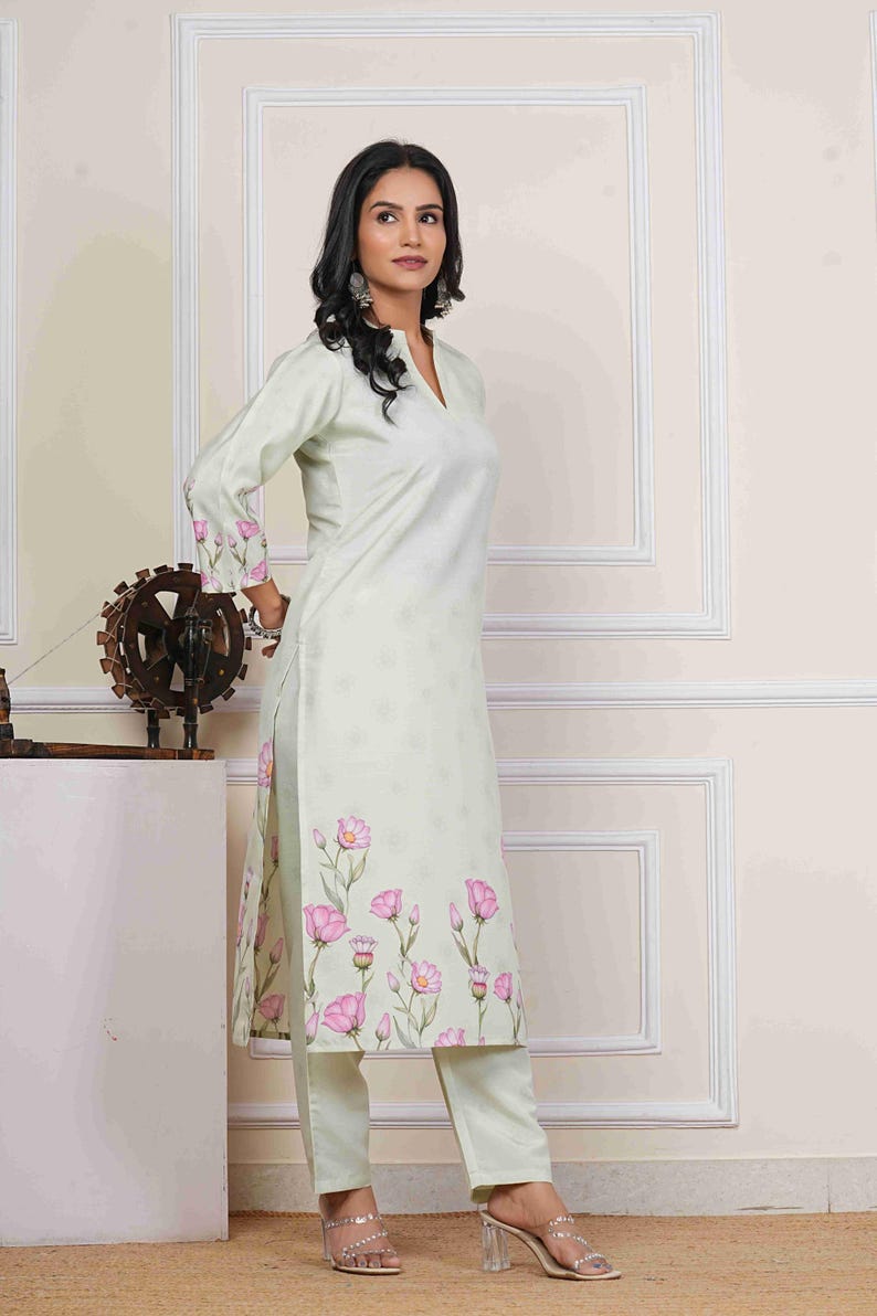 May include: A light green, long-sleeved kurta and matching pants set. The kurta features a V-neck and is adorned with a pink floral print along the hem and sleeves. The outfit is made of a lightweight fabric.