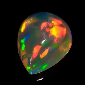 Puede incluir: Una piedra preciosa de ópalo en forma de pera con una vibrante exhibición de colores. El ópalo presenta destellos de rojo, naranja, amarillo, verde y azul, sobre una base blanca lechosa translúcida. La gema está sobre un fondo negro.