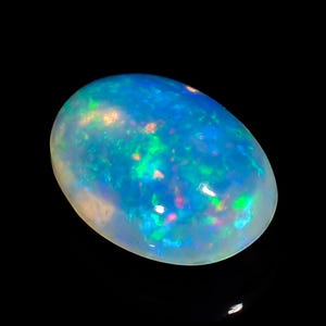 Può includere: Una gemma di opale lucidata, di forma ovale. La pietra mostra un vibrante gioco di colori, con lampi di blu, verde e accenni di rosso e giallo. L'opale è posto su uno sfondo nero, evidenziando le sue qualità iridescenti.