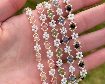 Op de afbeelding: Vijf kralenarmbanden met witte bloemvormige kralen en verschillende gekleurde middelste kralen. De armbanden zijn op elkaar gestapeld en worden in een hand gehouden.