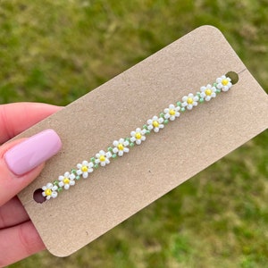 Op de afbeelding: Een witte en groene kralen armband met een madeliefje ontwerp. De armband ligt op een bruine kaart.