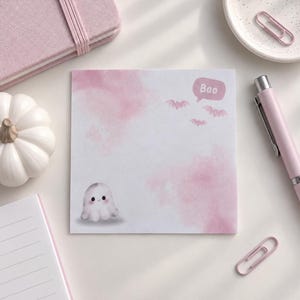 May include: A square notepad featuring a soft pink watercolour wash background, a small illustration of a smiling ghost in the bottom left corner, and three small bats near a speech bubble containing the text Boo in the upper right corner.