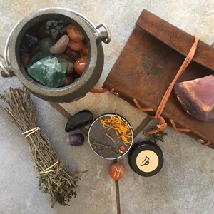 May include: A black cauldron filled with various crystals and stones, a bundle of dried herbs, a small black candle with orange wax, a black jar with a Capricorn symbol, a small red stone with the letter M, and a brown leather-bound journal with a stone on top.