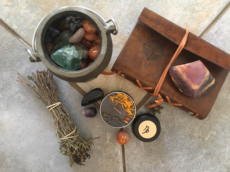 May include: A black cauldron filled with various crystals and stones, a bundle of dried herbs, a small black candle with orange wax, a black jar with a Capricorn symbol, a small red stone with the letter M, and a brown leather-bound journal with a stone on top.