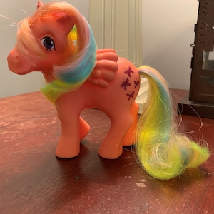 Puede incluir: Un juguete vintage de My Little Pony con un cuerpo rosa, una melena y una cola arcoíris y marcas de mariposa moradas.