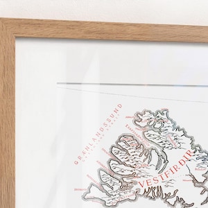 May include: A framed map print featuring the Westfjords region of Iceland. The map displays black outlines of the coastline and terrain with red text labels including Gr&aelig;nlandssund and Vestfir&eth;ir, all encased in a light wood frame.