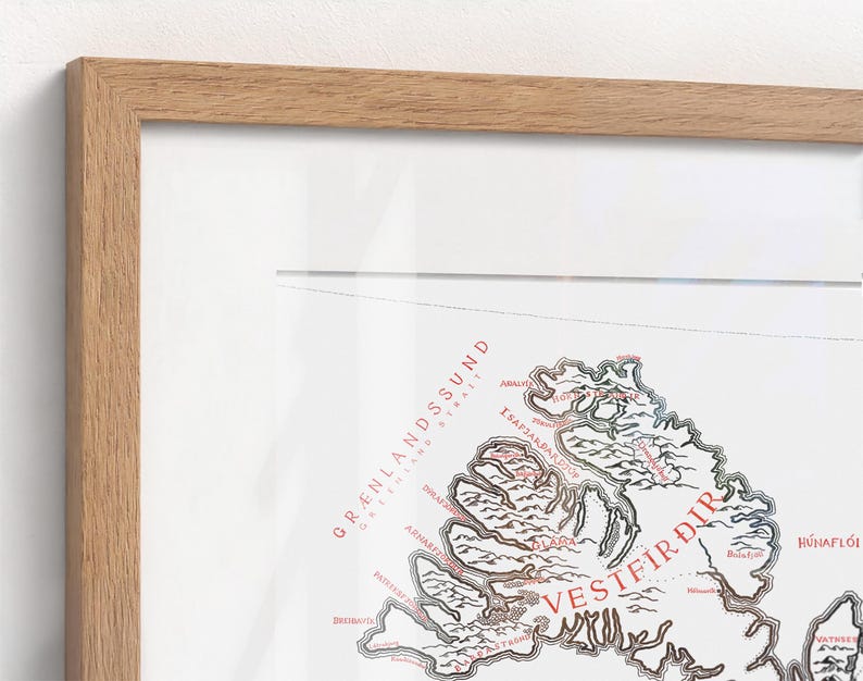 May include: A framed map print featuring the Westfjords region of Iceland. The map displays black outlines of the coastline and terrain with red text labels including Gr&aelig;nlandssund and Vestfir&eth;ir, all encased in a light wood frame.