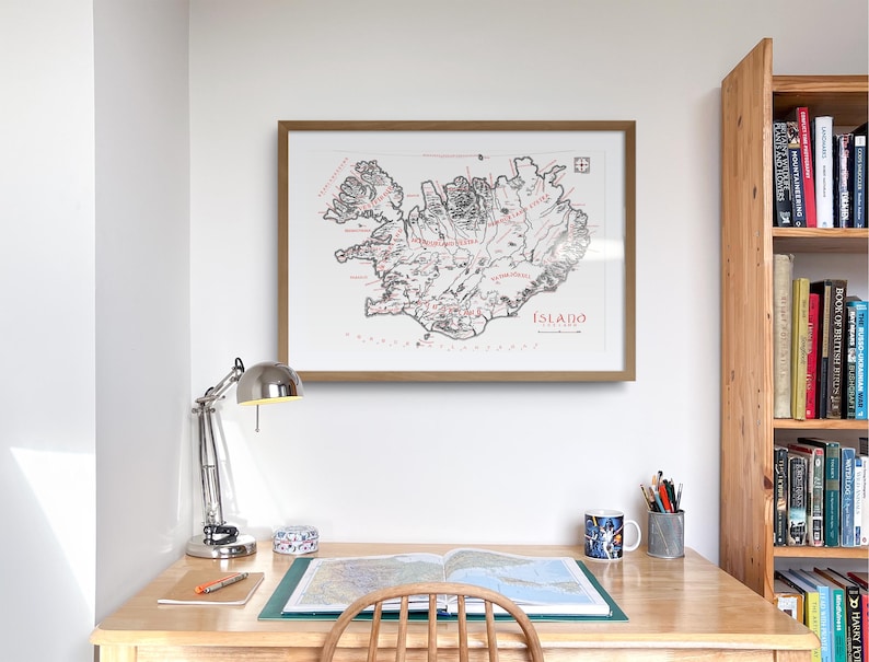 May include: A framed wall art print featuring a detailed black and white map of Iceland with red text labels, including the word &Iacute;SLAND. The map is displayed in a light brown wooden frame, showcasing geographical features and coastlines.