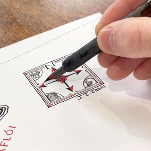 May include: A black ink pen held over a white paper featuring a hand-drawn square compass rose design with red and black accents. The illustration includes small corner sketches of a face, a helmet, and a structure, with the text J.T. Tabti written below.