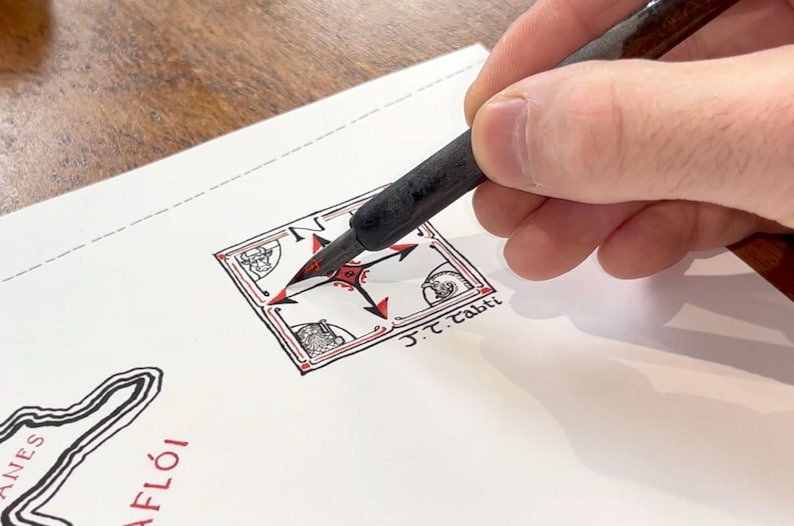 May include: A black ink pen held over a white paper featuring a hand-drawn square compass rose design with red and black accents. The illustration includes small corner sketches of a face, a helmet, and a structure, with the text J.T. Tabti written below.