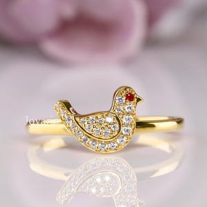 May include: A gold-colored ring featuring a bird design. The bird is encrusted with small, clear gemstones and has a red gemstone eye. The ring is set against a soft, blurred background.