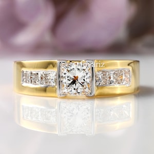 May include: A gold ring with a square, clear gemstone at the center, flanked by smaller rectangular gemstones. The ring has a gold band with a silver-colored setting around the center stone.