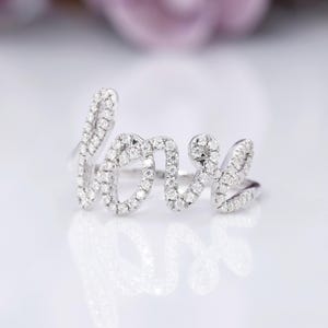 May include: A silver ring spelling out the word "love" in cursive script. The letters are encrusted with small, sparkling white gemstones. The ring is set against a white background, with a soft, blurred pink and purple backdrop.