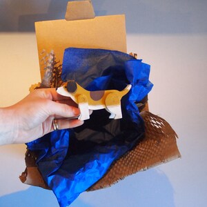 Pu&ograve; includere: Un cane giocattolo di legno dipinto di marrone, bianco e nero, &egrave; annidato in una scatola di cartone marrone foderata con carta velina blu. La scatola &egrave; piena di noccioline di imballaggio marroni.