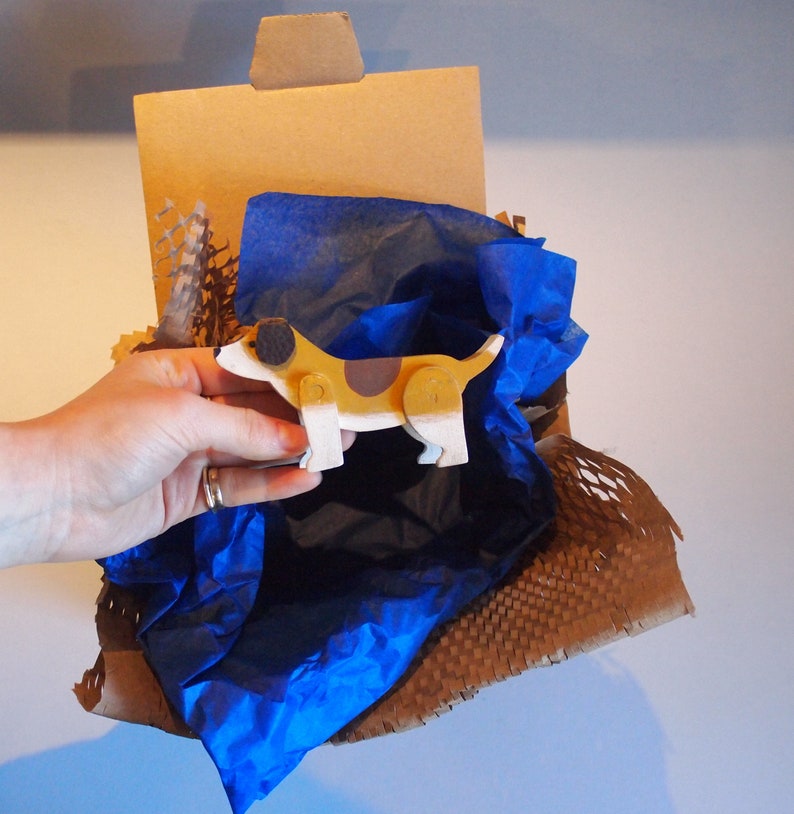 Pu&ograve; includere: Un cane giocattolo di legno dipinto di marrone, bianco e nero, &egrave; annidato in una scatola di cartone marrone foderata con carta velina blu. La scatola &egrave; piena di noccioline di imballaggio marroni.