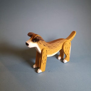Pu&ograve; includere: Un cane di legno giocattolo dipinto di marrone e bianco. Il cane ha una coda lunga e sta in piedi su tutte e quattro le zampe.