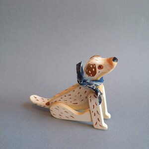 Pu&ograve; includere: Un cane giocattolo di legno con macchie marroni dipinte su uno sfondo bianco. Il cane indossa una bandana blu e bianca intorno al collo. Il cane &egrave; seduto con le zampe ripiegate sotto il corpo.