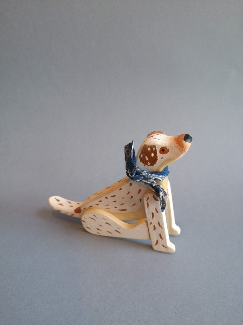 Pu&ograve; includere: Un cane giocattolo di legno con macchie marroni dipinte su uno sfondo bianco. Il cane indossa una bandana blu e bianca intorno al collo. Il cane &egrave; seduto con le zampe ripiegate sotto il corpo.