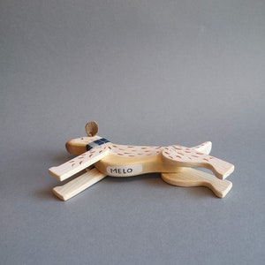 Pu&ograve; includere: Un cane giocattolo di legno con macchie marroni e bianche, che indossa una sciarpa blu e bianca. Il cane &egrave; sdraiato sul fianco con le zampe distese. Il cane ha un cartellino con il nome "MELO".