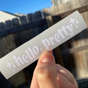 May include: A white rectangular sticker with the words "hello, pretty" in a stylized font. The word "pretty" is in a larger font size. White starburst graphics are on either side of the word "pretty".