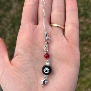 May include: A silver-colored charm necklace with a black eight ball bead, red and white beads, and a small silver star charm. The necklace is held in a hand against a blurred green background.