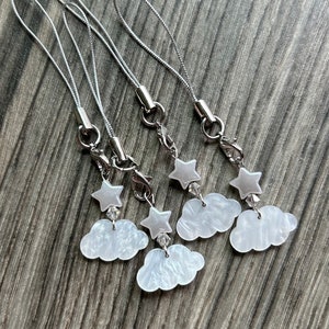 Puede incluir: Cinco dijes de nube blanca con una estrella plateada en la parte superior, cada uno unido a una cadena plateada con un cierre plateado.