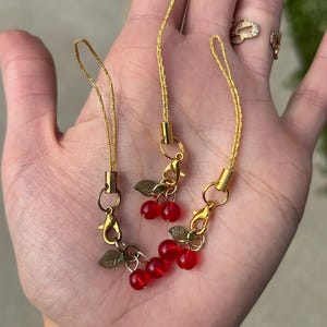 May include: Three gold cherry phone charms with red glass beads and green leaf accents. The charms are attached to gold cords.