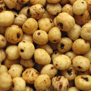 May include: A close-up of a bowl of light brown, round, puffed snacks. The snacks have a slightly rough texture and some have dark brown spots.