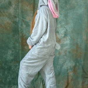 Op de afbeelding: Een grijs pluche konijnenkostuum met een roze binnenoor. Het kostuum heeft een capuchon en een lange staart. De persoon die het kostuum draagt, staat op een groene achtergrond.