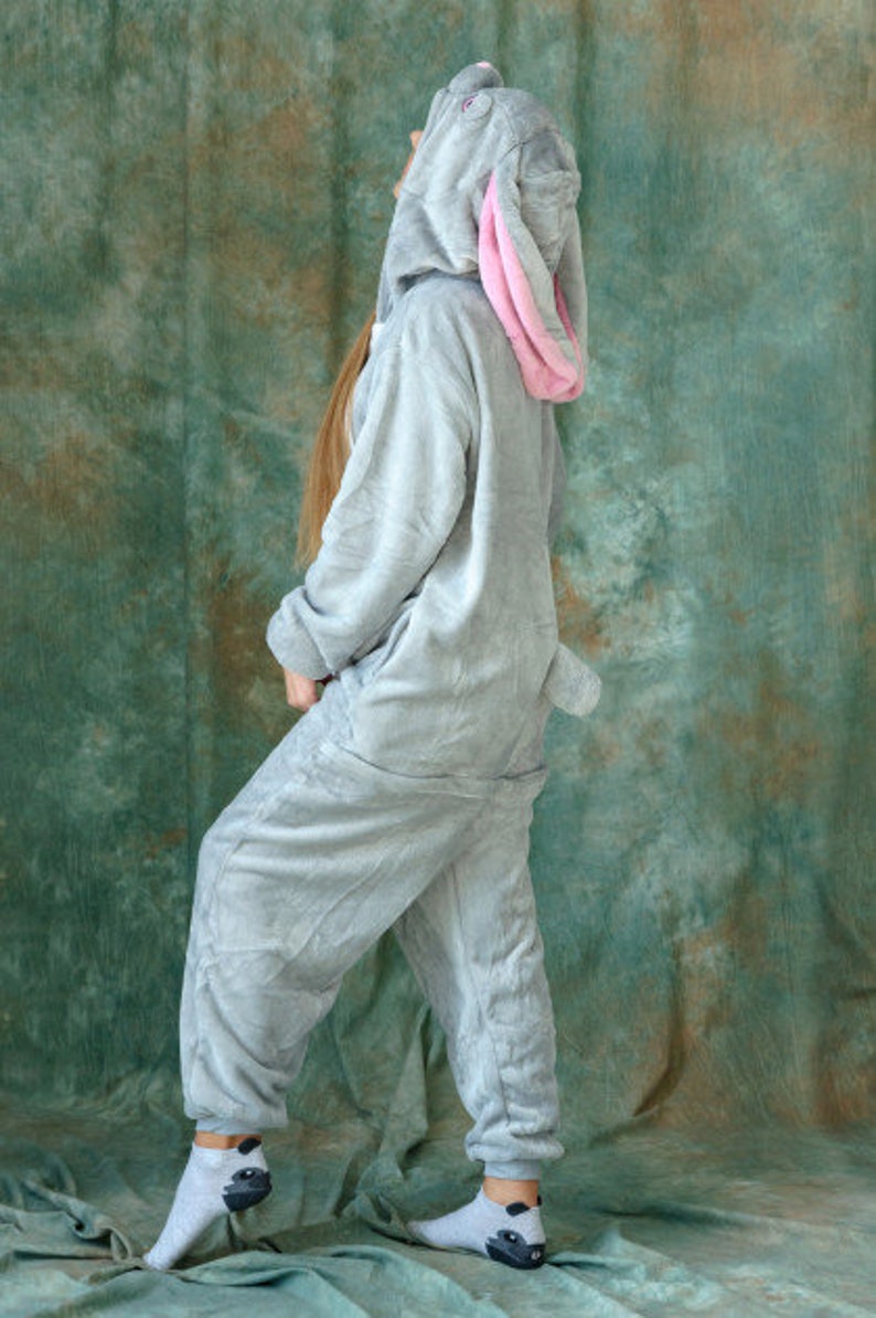 Op de afbeelding: Een grijs pluche konijnenkostuum met een roze binnenoor. Het kostuum heeft een capuchon en een lange staart. De persoon die het kostuum draagt, staat op een groene achtergrond.