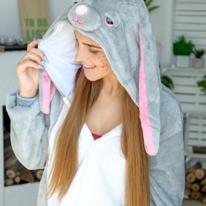 Op de afbeelding: Een vrouw draagt een grijze en roze bunny onesie met een witte voering. De onesie heeft een capuchon met bunny oren en een gezicht.