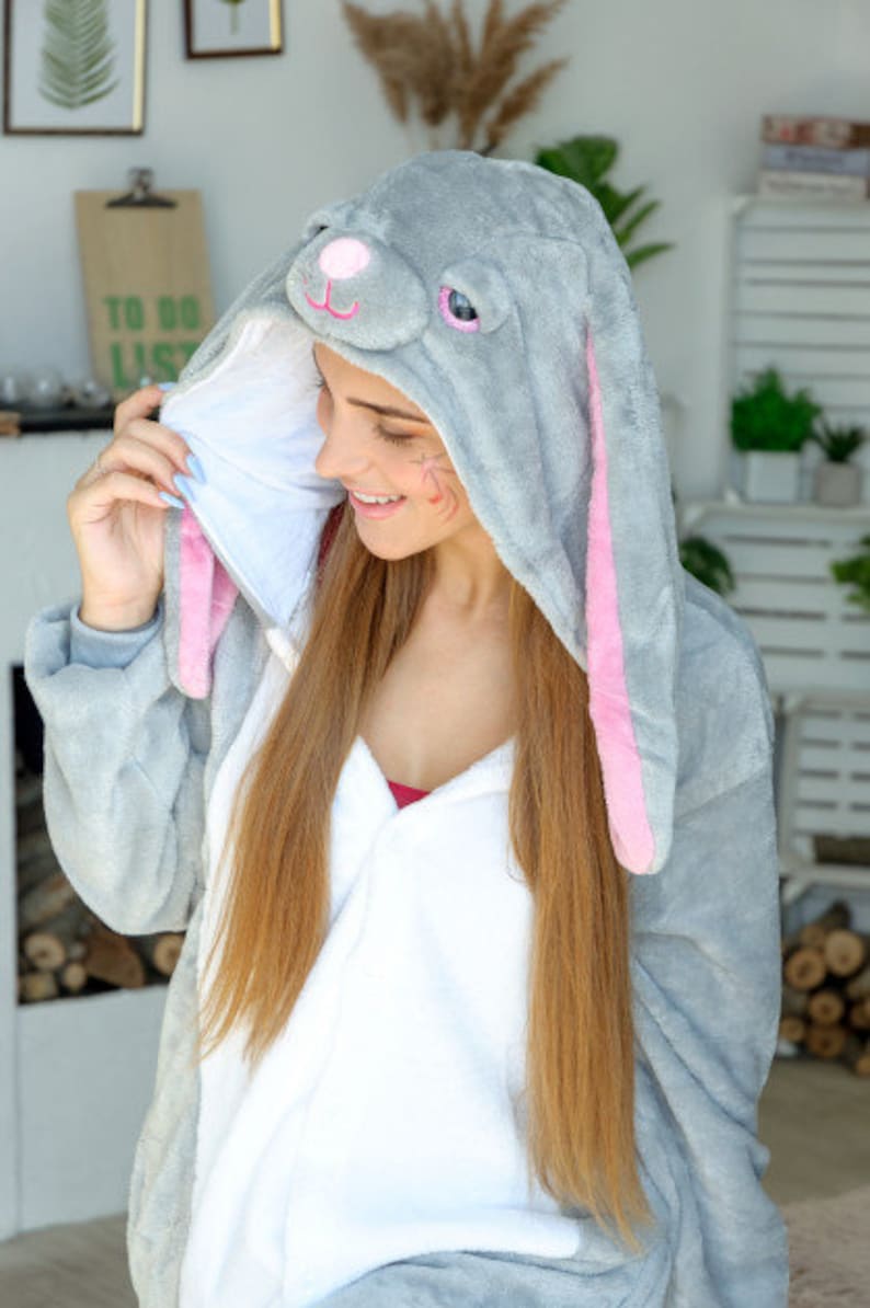 Op de afbeelding: Een vrouw draagt een grijze en roze bunny onesie met een witte voering. De onesie heeft een capuchon met bunny oren en een gezicht.