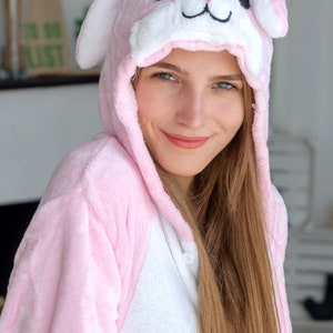 May include: A pink and white bunny onesie with a plush hood featuring a white bunny face with black eyes and a black nose.