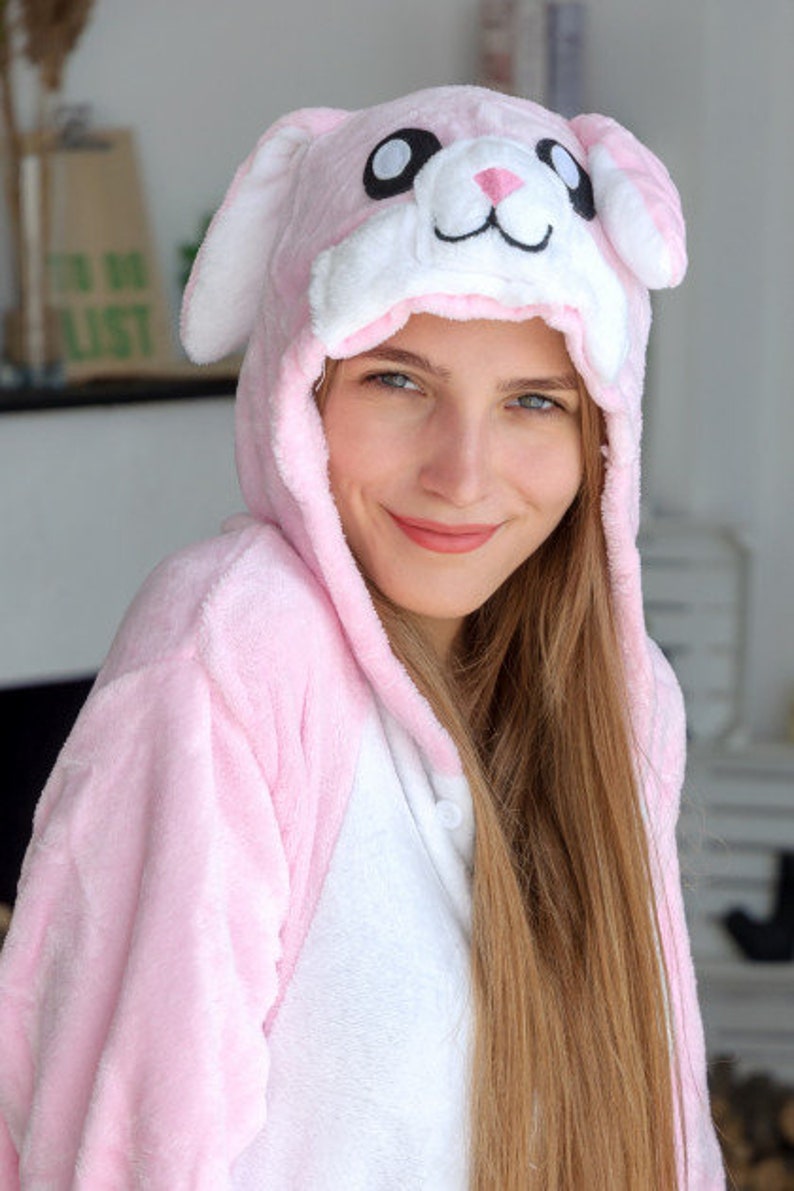 May include: A pink and white bunny onesie with a plush hood featuring a white bunny face with black eyes and a black nose.