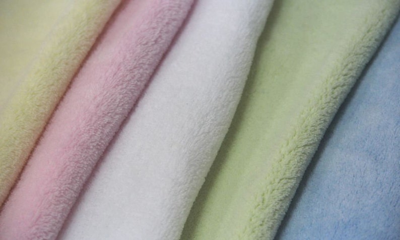 Op de afbeelding: Vijf stofstalen in verschillende kleuren: lichtgeel, roze, wit, lichtgroen en lichtblauw. De stof is zacht en pluche, met een pluche textuur.