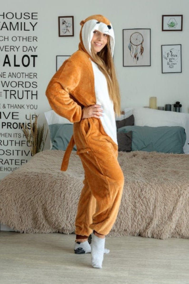 May include: A woman wearing a brown and white animal onesie with a hood. The onesie has a long tail and the hood has a cute animal face. The woman is standing on a light brown rug.
