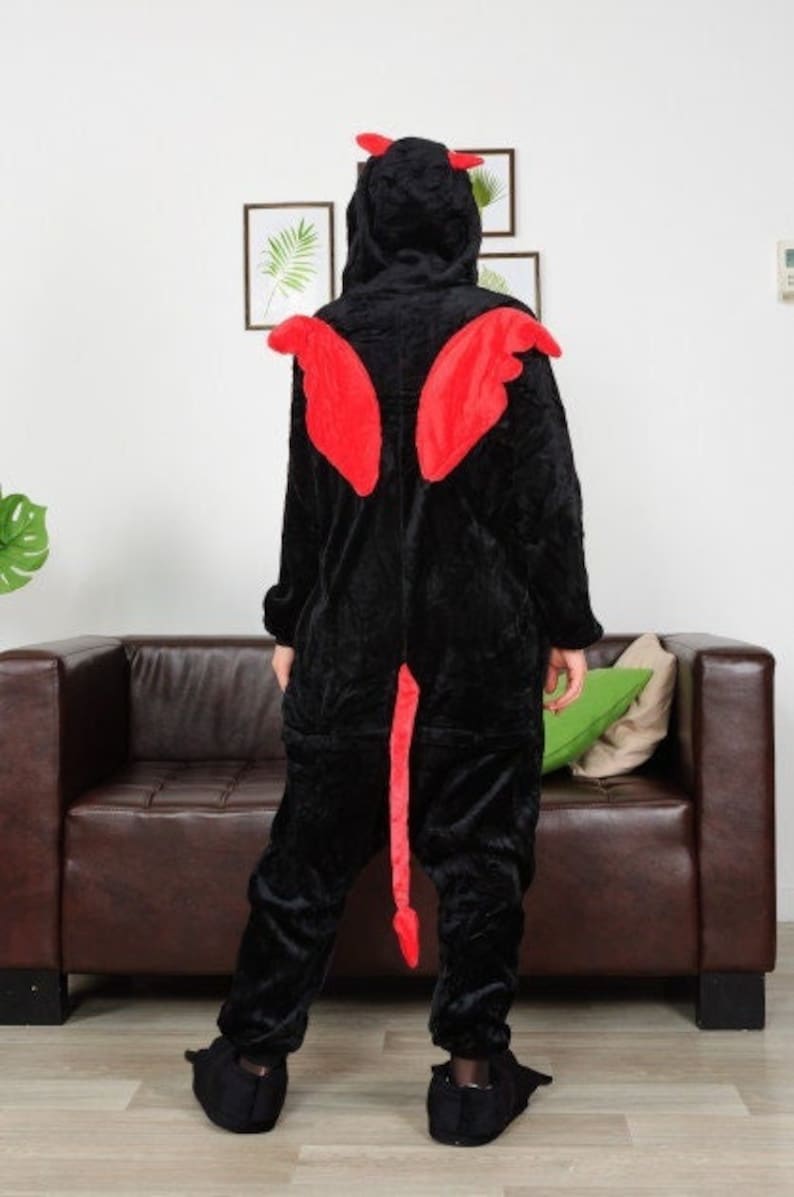 Op de afbeelding: Zwarte fleece onesie met een rode duivelsstaart, hoorns en vleugels. De onesie heeft een capuchon en wordt door iemand gedragen.