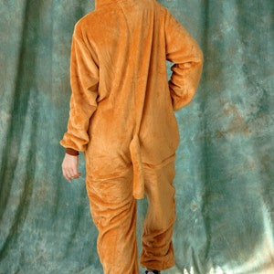 May include: A brown hooded jumpsuit with a white hood lining. The jumpsuit has a long tail and is made of a soft, plush material.