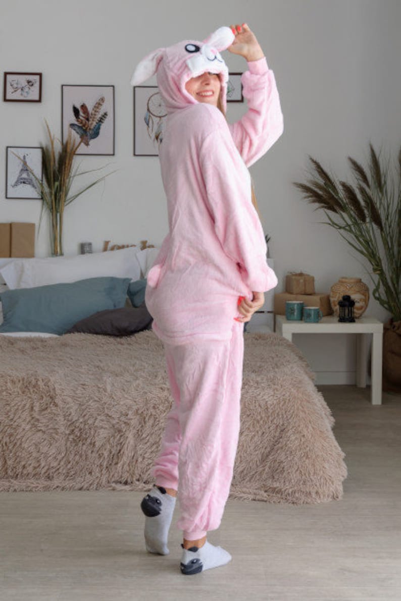 May include: A pink bunny onesie with a white bunny face on the hood. The onesie has long sleeves and long pants. The person wearing the onesie is standing on a fluffy rug.