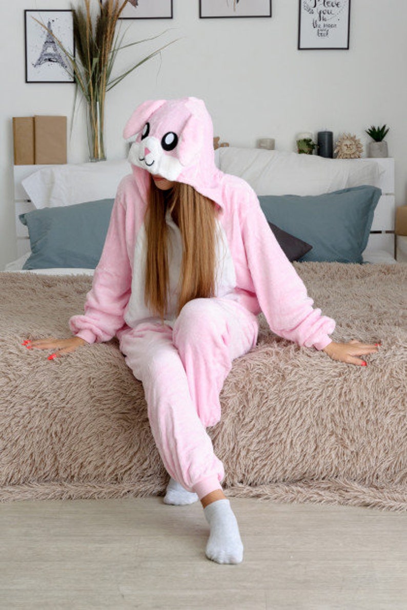 May include: A pink bunny onesie with a white belly and a hood featuring a bunny face. The onesie is worn by a person sitting on a fluffy, light brown rug.