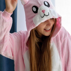 May include: A pink bunny onesie with a white belly and a white face with black eyes. The onesie has a hood with bunny ears.