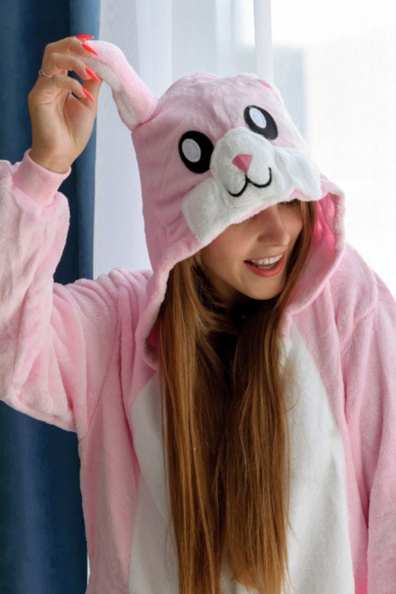 May include: A pink bunny onesie with a white belly and a white face with black eyes. The onesie has a hood with bunny ears.