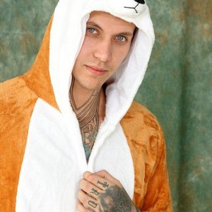 May include: A person wearing a brown and white animal onesie with a hood shaped like an animal head. The onesie has a zipper down the front and the hood has a white fur lining. The person has tattoos on their arms and hands.