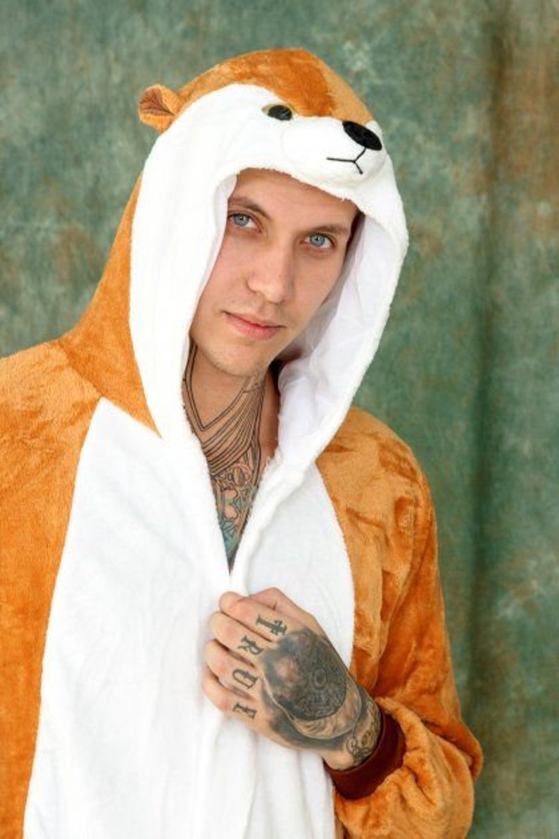 May include: A person wearing a brown and white animal onesie with a hood shaped like an animal head. The onesie has a zipper down the front and the hood has a white fur lining. The person has tattoos on their arms and hands.