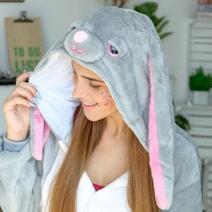 Op de afbeelding: Een grijze en roze bunny onesie met lange mouwen. De capuchon heeft een pluche konijnengezicht met roze oren en zwarte ogen.