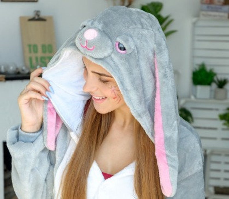 Op de afbeelding: Een grijze en roze bunny onesie met lange mouwen. De capuchon heeft een pluche konijnengezicht met roze oren en zwarte ogen.