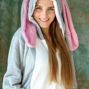 Op de afbeelding: Een vrouw draagt een grijze en roze bunny onesie met een witte voering. De onesie heeft een capuchon met bunny oren en een schattig gezicht.